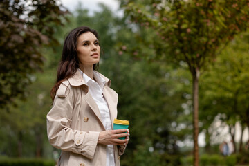 A girl brunette walks in the spring park and holds a cup of coffee in hands. A lady with serious gaze looks into the distance thoughtfully. A young woman enjoys the beauty and harmony of nature alone