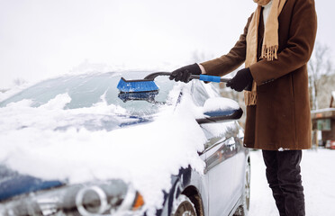 Young man in a stylish coat cleans the car from snow with a brush. Winter glass washing. Clearing snow from a car. Seasonality concept.