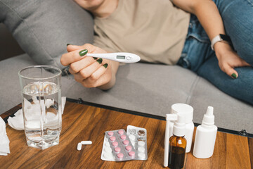 Sick woman with fever lying on sofa at home, holding digital thermometer. cold remedies, medicine and pills on table. Concept of seasonal diseases, coronavirus and flu