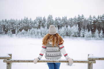 Beautiful young woman in winter clothes enjoying the winter forest. Behind her is a snow-covered lake. Happy winter time. Christmas.