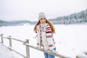 Beautiful young woman in winter clothes enjoying the winter forest. Behind her is a snow-covered lake. Happy winter time. Christmas.