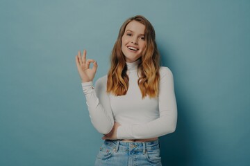 Smiling woman making OK sign with hand, dressed in white turtleneck and jeans on a blue background.