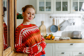 portrait of smiling young blonde woman in red sweater. housewife stands near cupboard in red sweater in cozy kitchen during morning