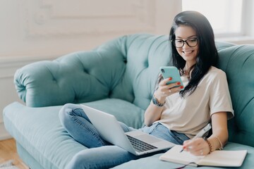 Smiling student girl working on laptop with smartphone and notebook on a comfy blue sofa at home