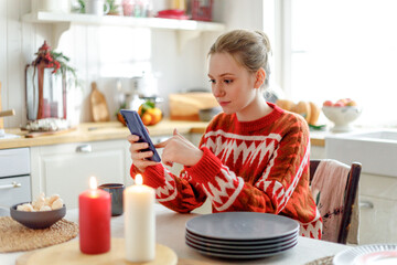 young blonde pensive woman in red sweater sits in cozy kitchen with Christmas decorations. looks calm in smartphone screen
