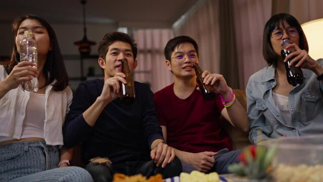 Group of Asian people friends sit on sofa watching and cheering football or soccer games competition on TV together at home.Happy man and woman sport fans celebrating sport team victory sports match