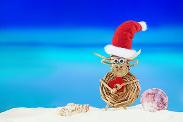 Toy woven cow with Santa' hat with heart and shells on sandy beach with sea. Christmas, New Year,...