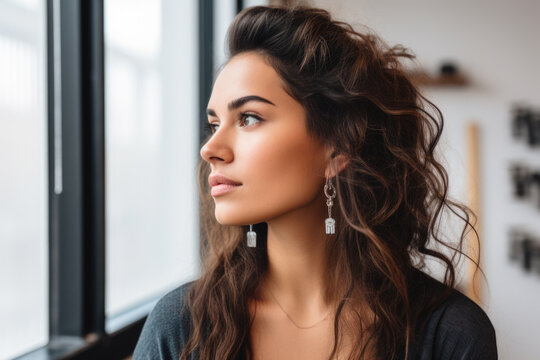 Woman is seen looking out of window while wearing earrings. This image can be used to depict curiosity, anticipation, or introspection. It is suitable for various projects and designs