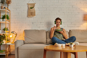 delighted woman in headphones having vegetarian dinner on couch in cozy living room, quiet evening