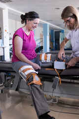 Vertical photo of a middle aged female patient sitting in front of her physiotherapist as she puts the prosthesis on her leg.