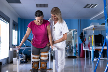 A middle aged female patient with prosthesis in both legs is walking with the help of her physiotherapist on the parallel bars of the hospital gym. - Powered by Adobe