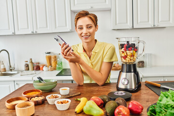 vegetarian woman with smartphone smiling at camera near plant origin food and blender with fruits