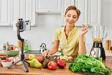 cheerful vegetarian woman looking at smartphone on tripod near fruits and electric blender, vlog