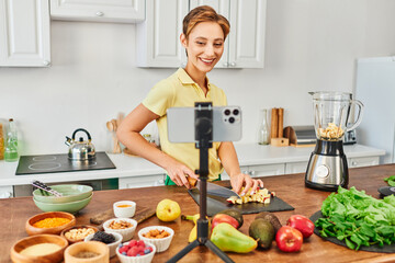 happy vegetarian woman cutting fruits near blender and smartphone on tripod, plant-based diets vlog