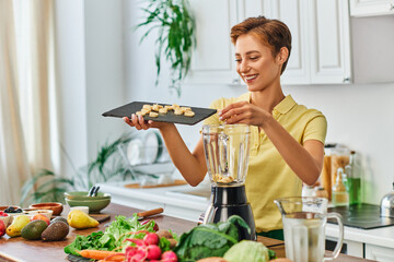 joyful woman with sliced banana on chopping board near blender and vegetarian ingredients in kitchen