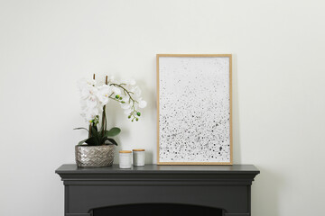 Picture frame, candles and orchid on fireplace near white wall indoors. Interior element