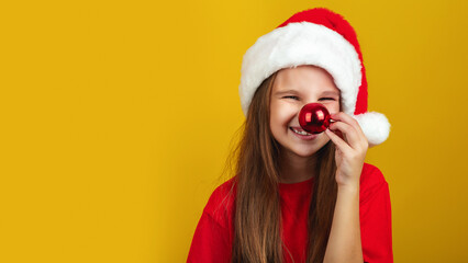Holiday Fun. Cheerful Little Girl Wearing Santa hat, Playful Kid Keeping Xmas Tree Ball Near Nose And Looking At Camera, Yellow Background. Copy Space