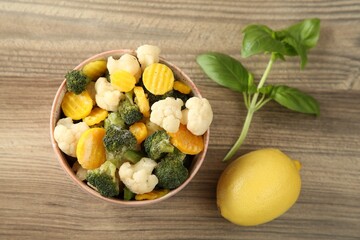 Frozen vegetables in bowl, basil and lemon on wooden table, flat lay