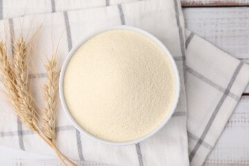 Uncooked organic semolina in bowl and spikelets on white wooden table, top view