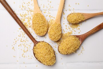 Spoons with raw bulgur on white tiled table, flat lay
