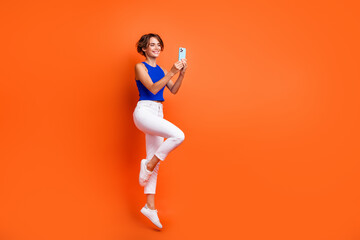 Full body profile photo of lovely active lady jump use smart phone texting empty space isolated on orange color background