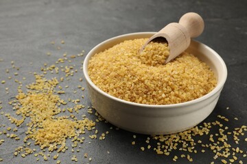Bowl and scoop with raw bulgur on dark gray table, closeup