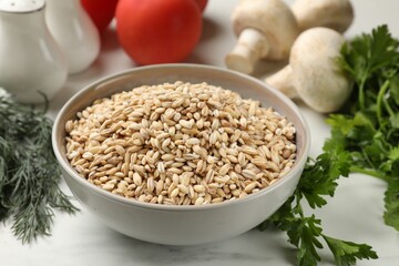 Dry pearl barley in bowl and products on white marble table