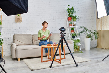 video blogger with smartphone near vegetarian food and digital camera with softbox lamp at home