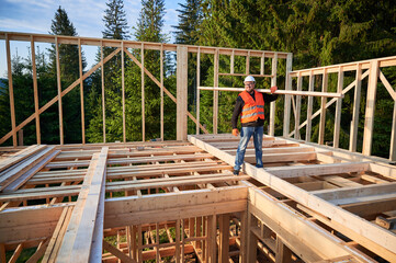 Carpenter builds wooden frame house near woods. Man holds large beam on shoulder, wearing work clothes and helmet. Concept of modern and sustainable building.