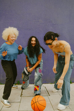 Three multiracial friends in fashionable outfits playing basketball in front of blue wall outdoors