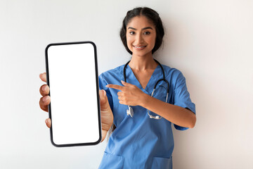 Indian Doctor Woman Wearing Blue Scrubs Pointing At Blank Smartphone In Hand