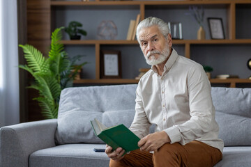 Portrait of a gray-haired senior man sitting at home on the couch, holding a book in his hands and looking seriously into the camera