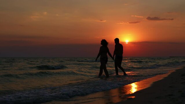 A man and a woman walk into the sea holding hands against the backdrop of a beautiful sunset. Slow Motion