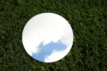 Round mirror on green grass reflecting cloudy sky, top view