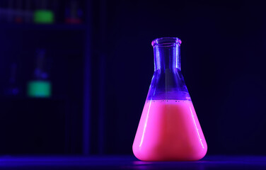 Laboratory flask with luminous liquid on table against dark background, space for text