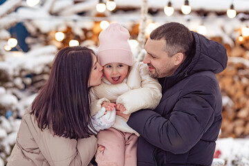 Happy family mom, dad and little daughter on Christmas market outdoor. A beautiful young family stands on a street with festive Christmas decoration. Snow, Christmas. Happy holidays.