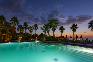 Dawn at the basem - a pool with clear illuminated water and palm trees growing on the edges in the...