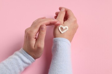 Woman with heart made of cosmetic cream on hand against pink background, closeup
