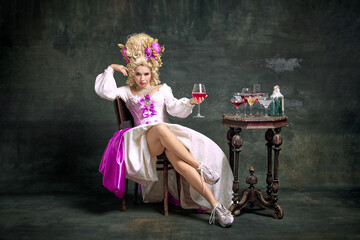 Serious princess, aristocratic medieval person in old-fashioned dress and modern shoes sitting holding cocktail against vintage background. - obrazy, fototapety, plakaty
