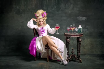 Fotobehang Serious princess, aristocratic medieval person in old-fashioned dress and modern shoes sitting holding cocktail against vintage background. © Lustre