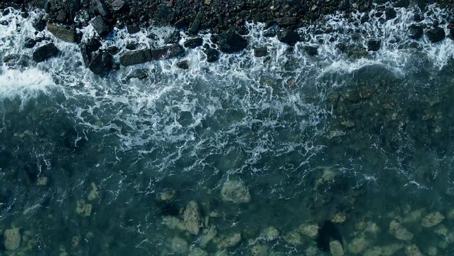 Waves of sea break on the rocky shore. Transparent clean water and the seabed near the shore. Aerial top down shot