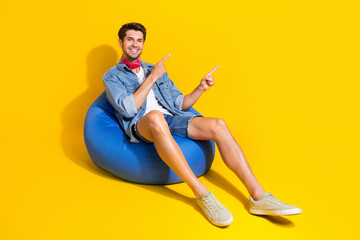 Full size photo of positive guy wear jeans jacket shorts indicating at logo empty space sit on pouf isolated on yellow color background