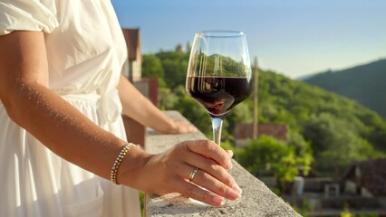 Elegant brunette woman, holding a glass of red wine, steps out onto the balcony or villa terrace and gazes at the mesmerizing sunset over the mountains. Travel, summertime, and the beauty of nature