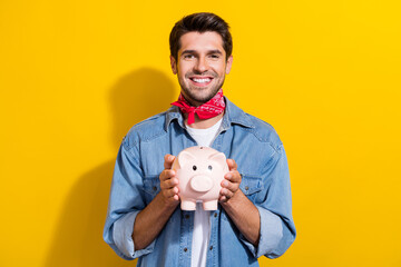 Fototapeta na wymiar Photo portrait of nice young man hold piggy account toothy smile wear trendy jeans garment isolated on yellow color background