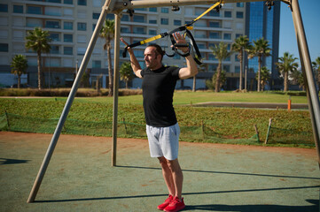 Young adult sportsman doing bodyweight exercises on arms, push ups with suspension straps in urban...