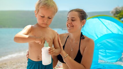 Portrait of little baby boy with mother applying sunscreen UV protection lotion on sea beach during...