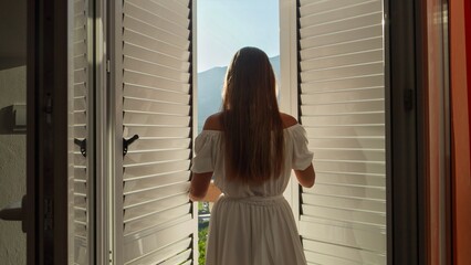 Young brunette woman in long dress opens balcony doors, walks out on the terrace and enjoys sunrise or sunset over the mountains. Concept of summertime holiday, vacation, enjoying nature and travel