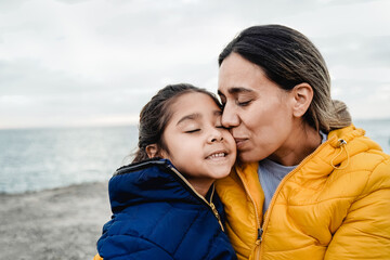 Happy Latina mother and child daughter hugging each other in winter time with background beach. Family love and travel vacations - obrazy, fototapety, plakaty