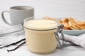Tasty condensed milk in jar, crepes and vanilla pods on white wooden table, closeup