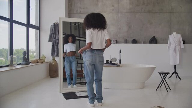 Black Woman in Big Jeans Standing in Bathroom. Cheerful African American female after successful weight loss. Slim fit woman in bathroom.